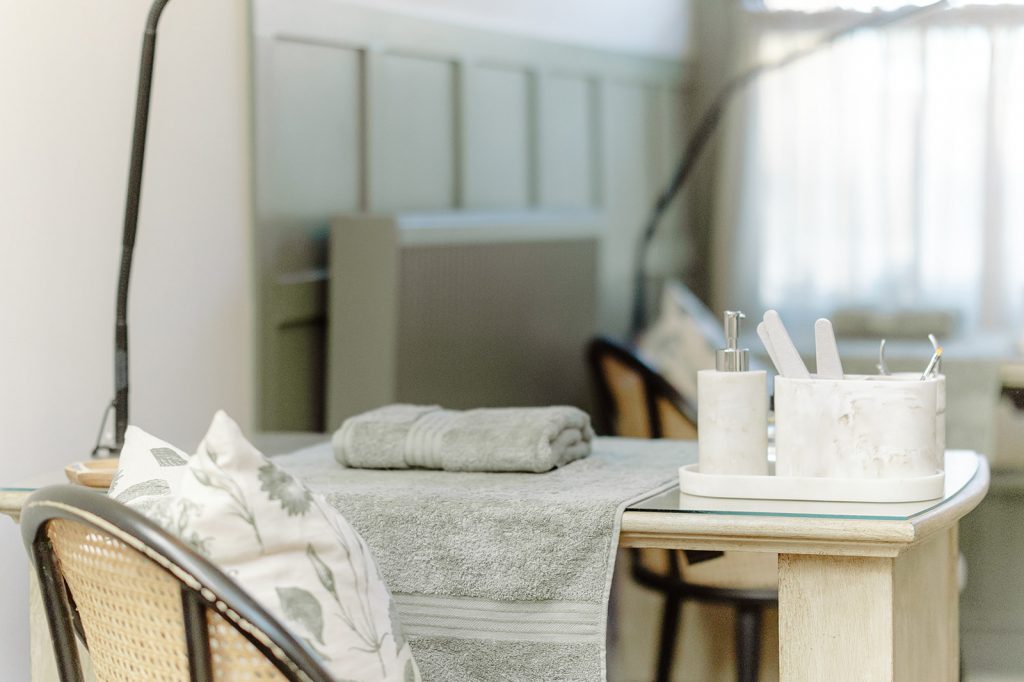 A therapy room with a nail desk and a rattan chair tucked under, towels folded on desk and sage green panelling on the wall